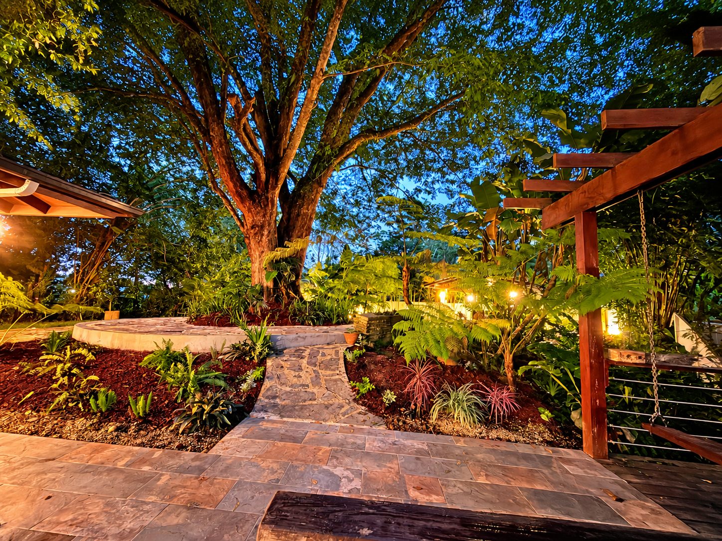 Jardín de Hacienda Rosaura al atardecer, con un árbol grande, un camino de piedra y luces encendidas